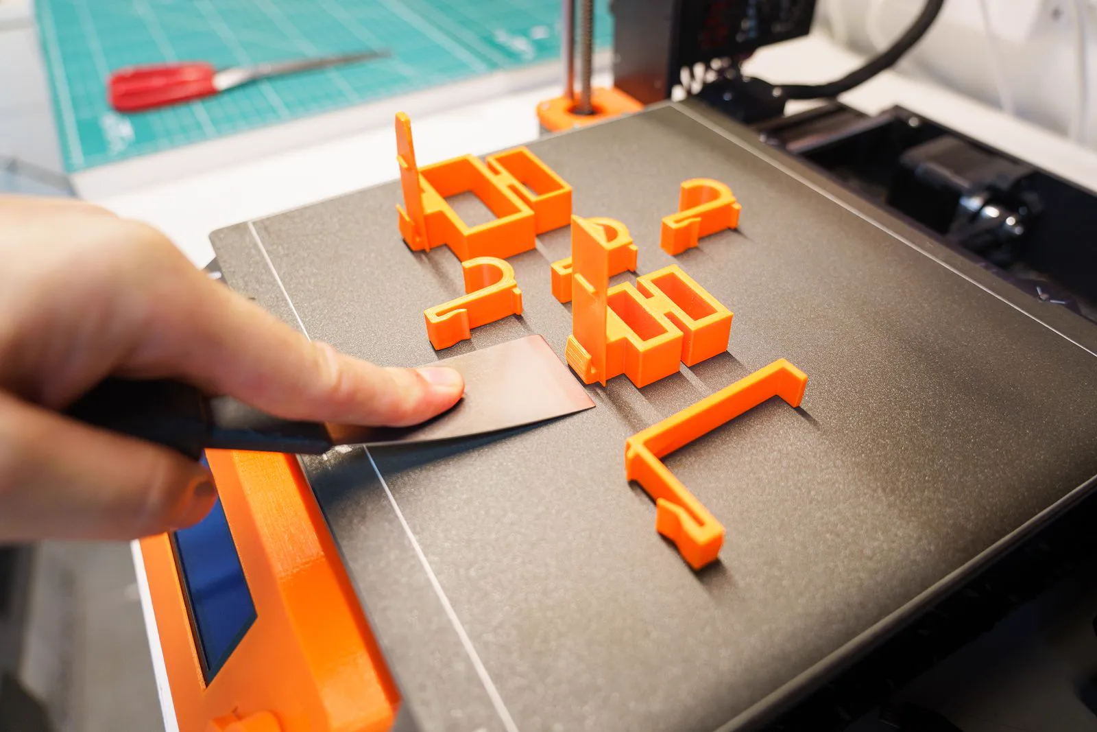 Mão retirando uma peça laranja recém-impressa da mesa de uma impressora 3D com uma espátula
